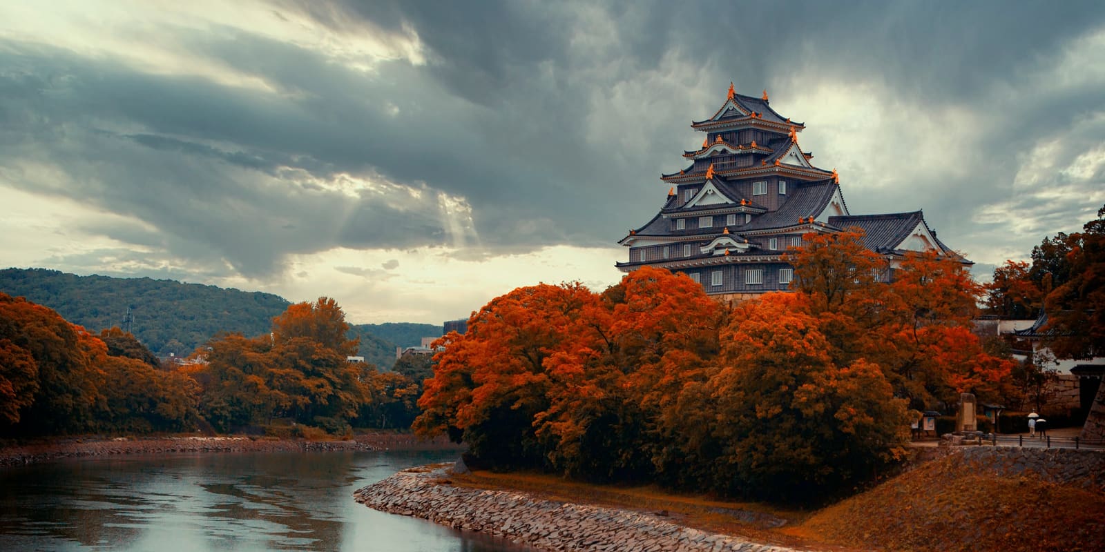A black castle keep with gold trim stands above a riverbank of red and orange trees, under heavy grey clouds