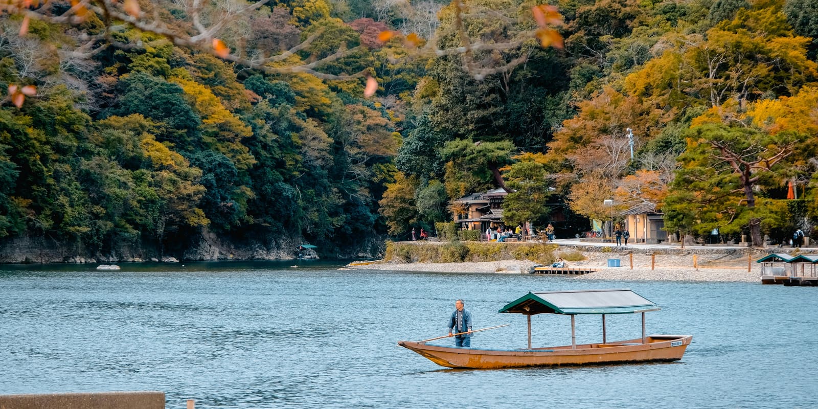 A boatman stands and poles a covered wooden boat across a calm river, below a wooded hillside turning orange and gold