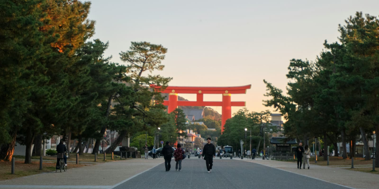 A giant vermilion torii gate at the end of a wide, pine-lined avenue at dusk, with people walking and cycling toward the camera