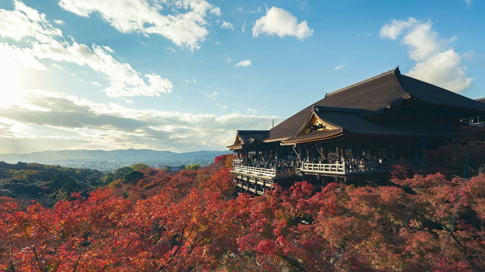 Kiyomizu-dera's Main Hall and wooden stage above red autumn maples, with Kyoto and the western mountains beyond