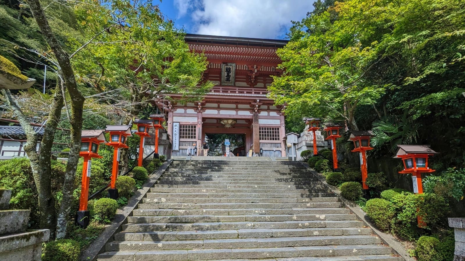 Stone steps lined with red lanterns leading up to Kurama-dera's vermilion Niomon gate, among green maples