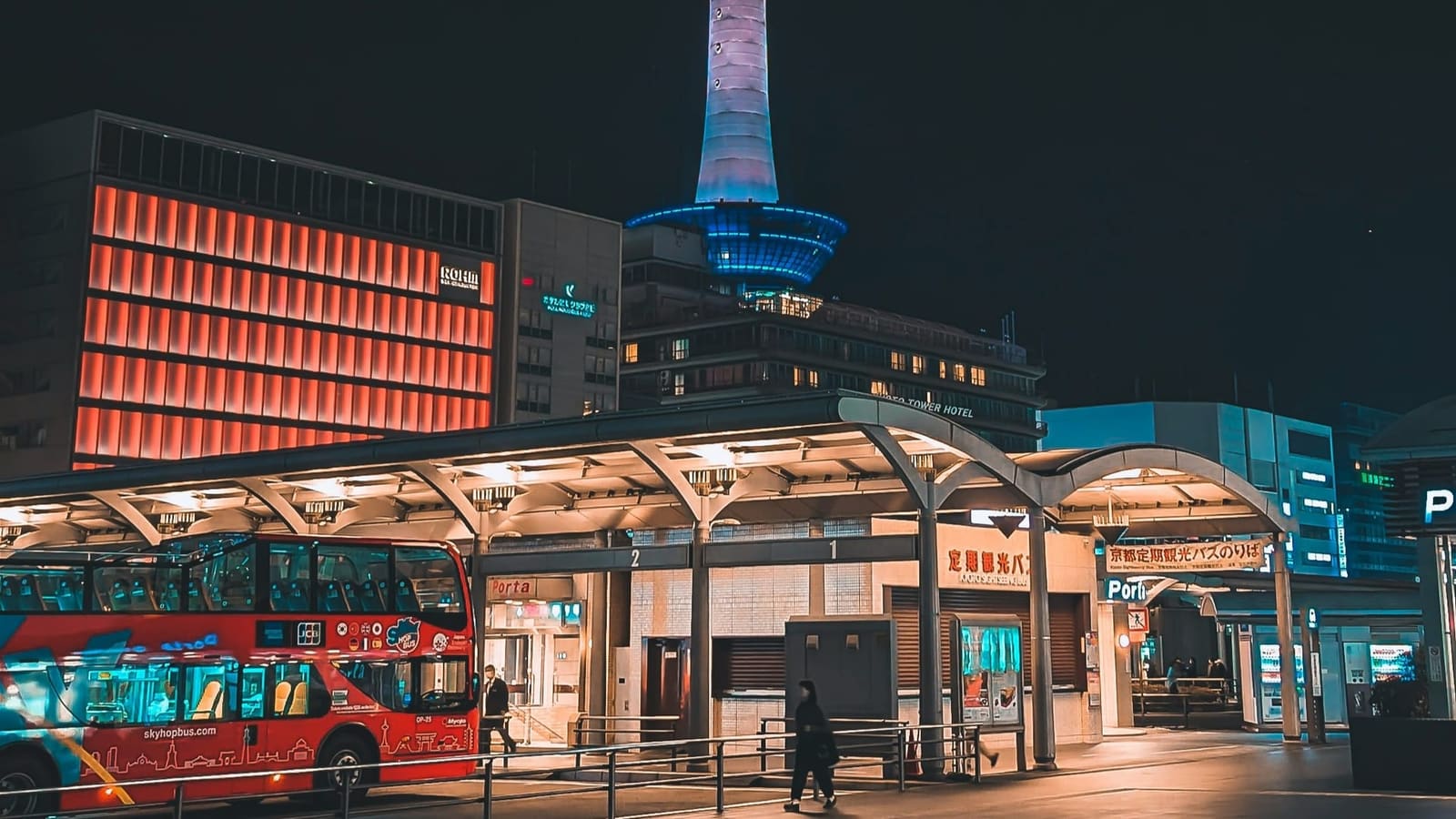 Kyoto Tower lit blue at night above the Kyoto Station bus terminal, with a red open-top sightseeing bus at the stop