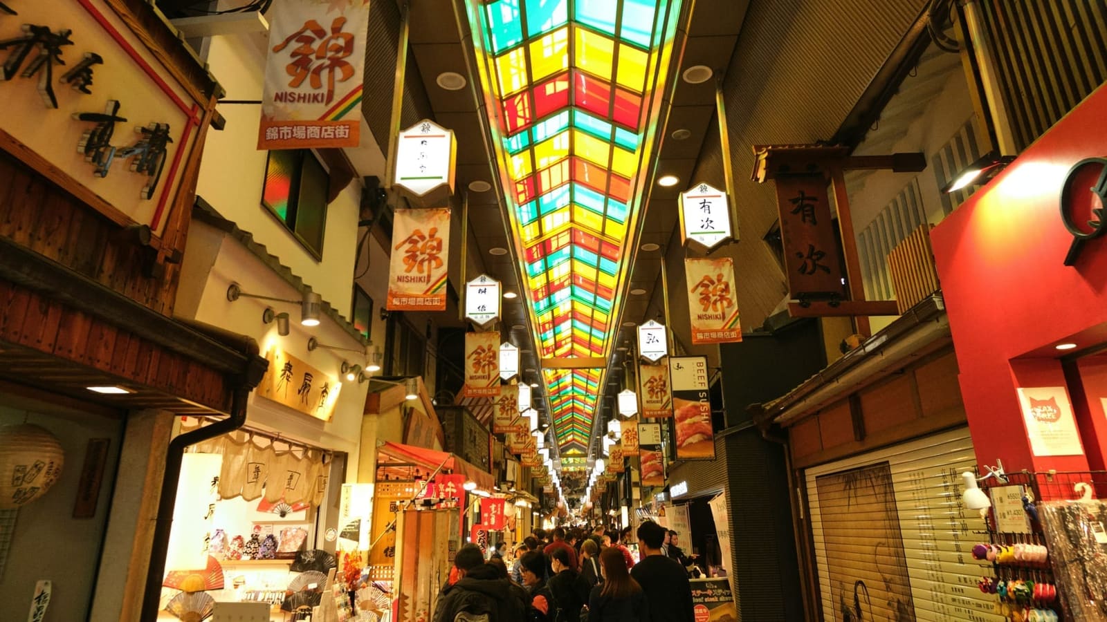 The covered Nishiki Market arcade in Kyoto, with its red, yellow and green glass roof and hanging Nishiki banners above shoppers
