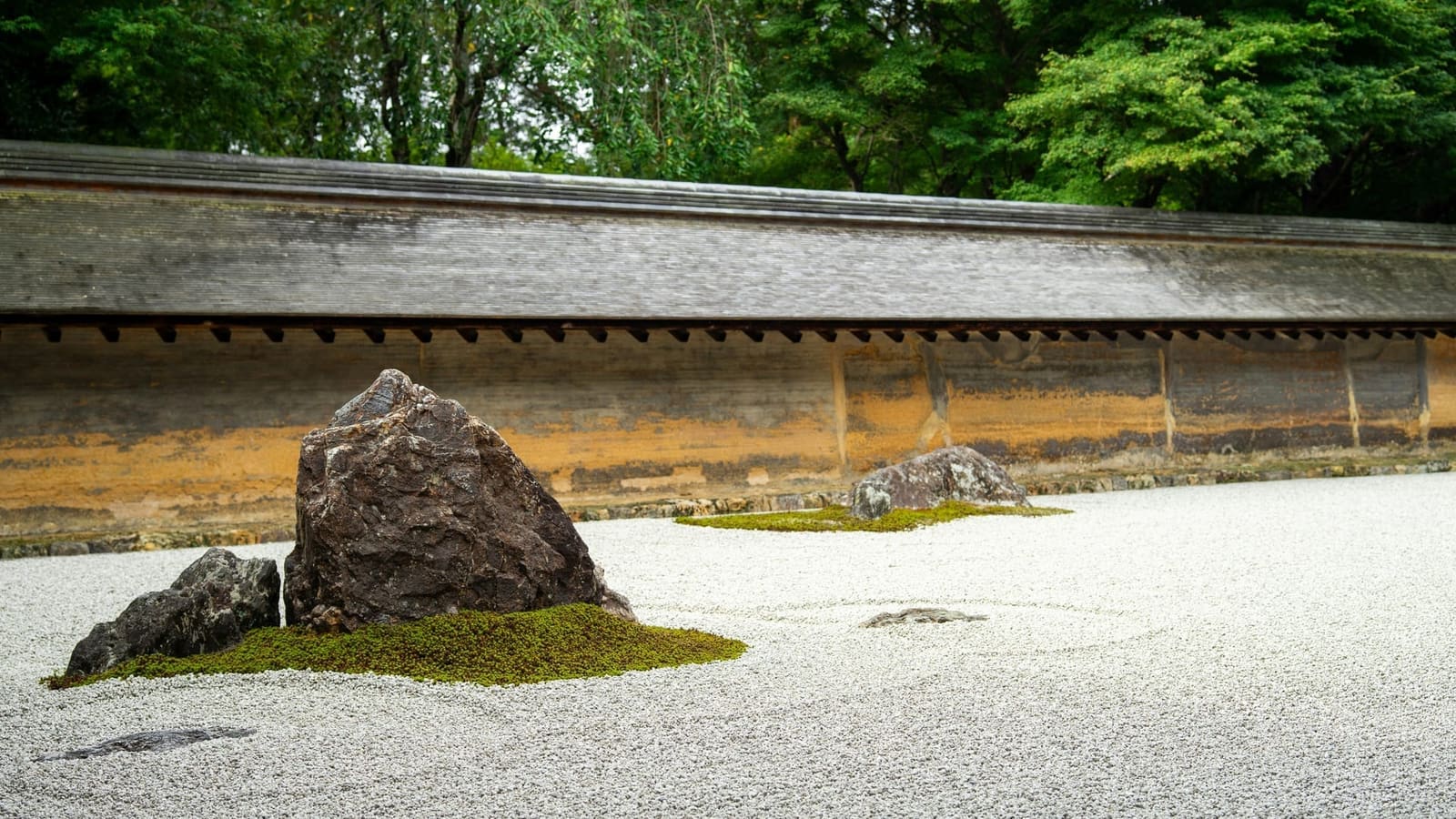 A stone group on moss in Ryoan-ji's raked gravel, in front of the weathered oil-clay wall