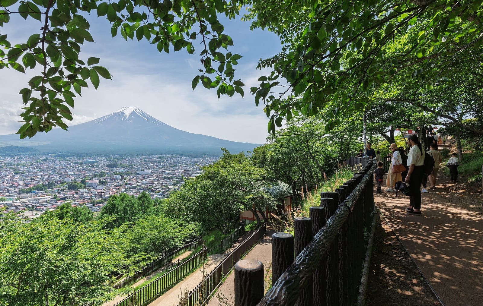 Hillside footpath with wooden railings in Arakurayama Sengen Park, visitors walking under green trees, with the town of Fujiyoshida and Mount Fuji beyond