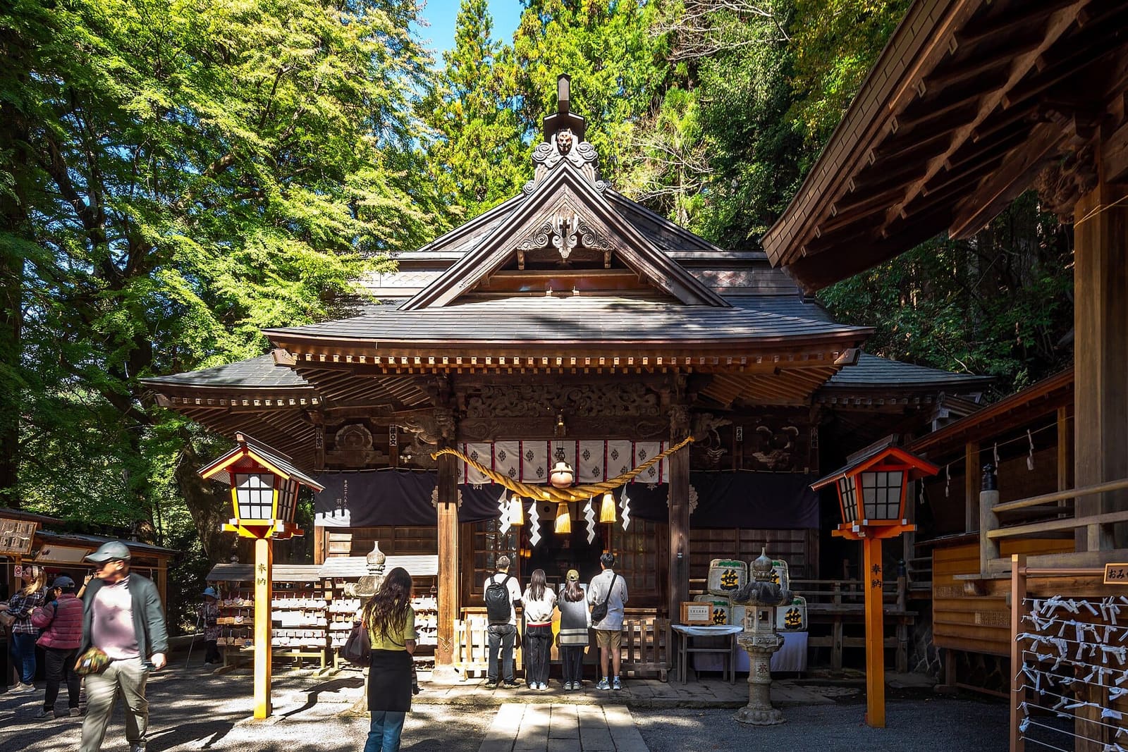Hall of Arakura Fuji Sengen Shrine with a thick straw rope and paper streamers, orange lanterns on either side and visitors praying in front, among tall trees