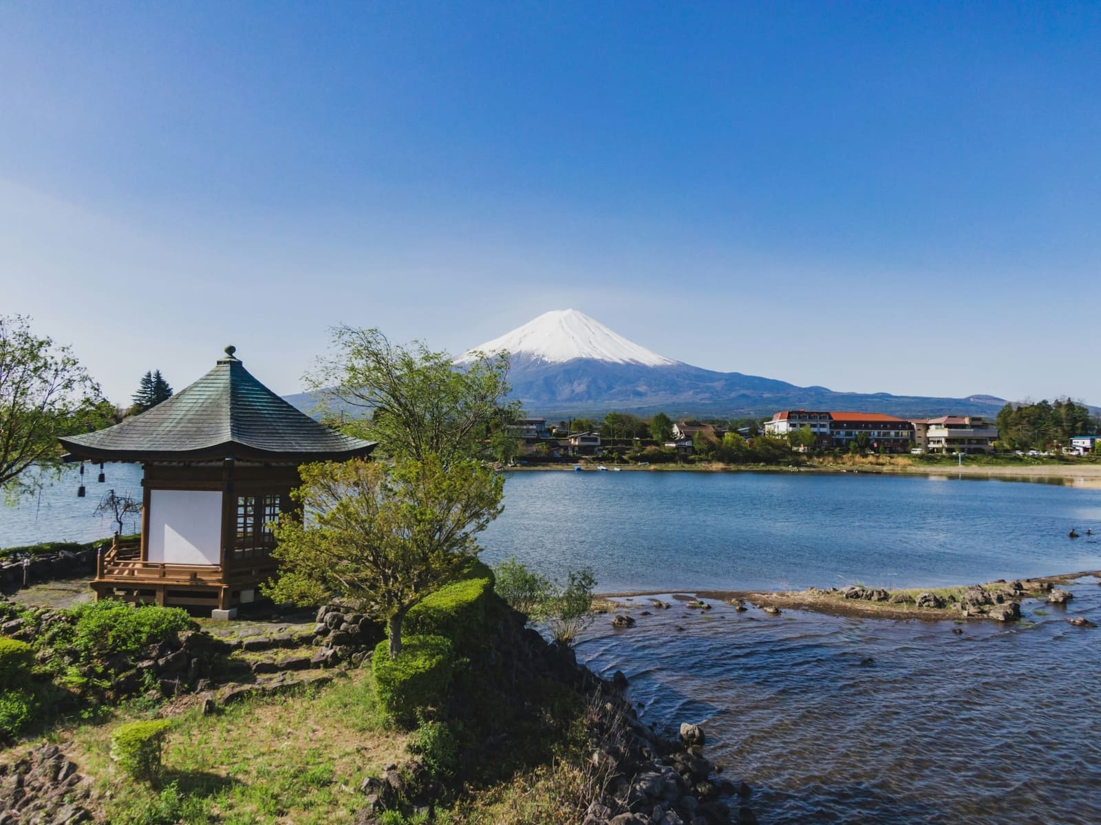 Small lakeside pavilion on rocks at Lake Kawaguchiko with snow-capped Mount Fuji across the water