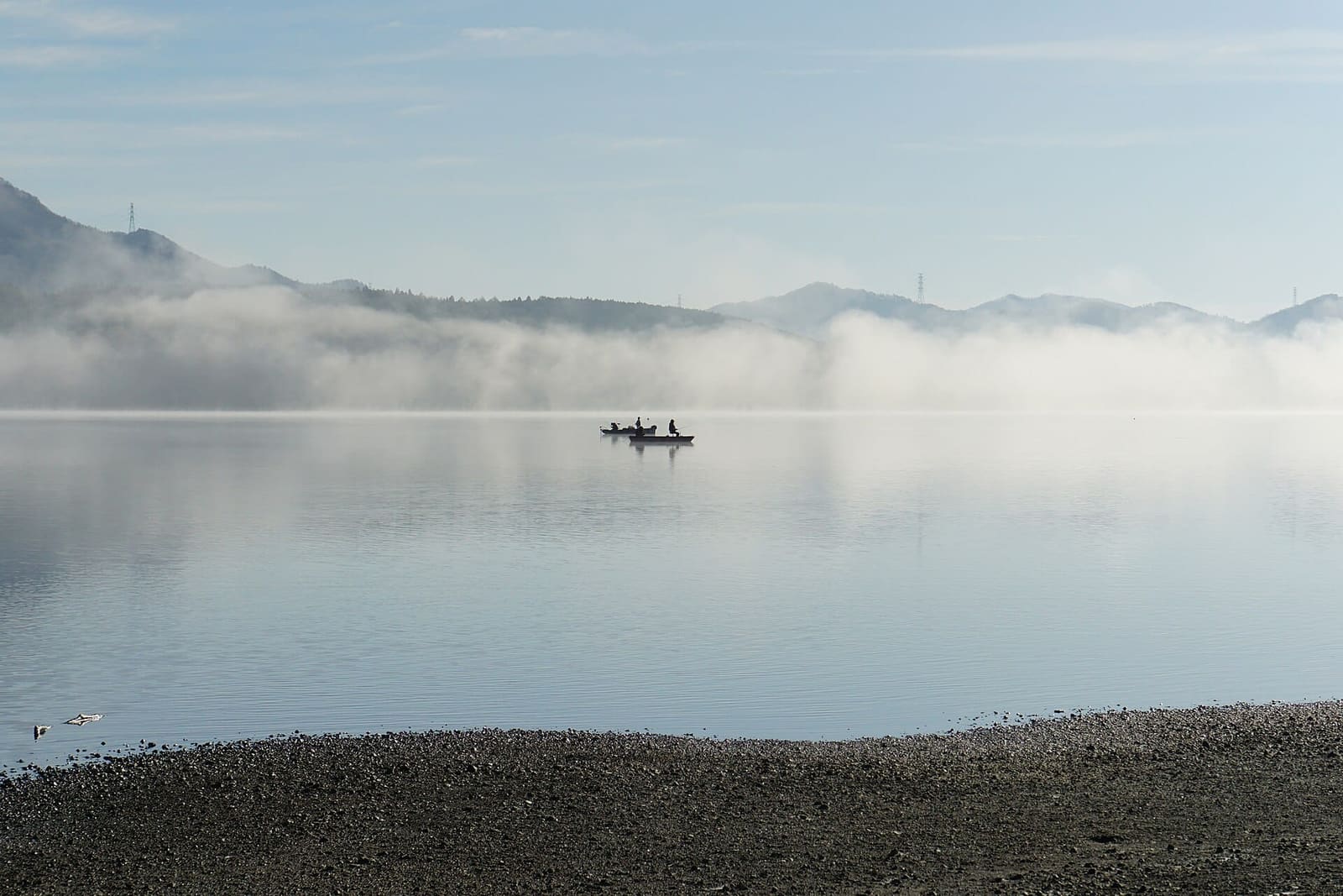 Small fishing boats on Lake Yamanakako in early-morning mist, with a stony shore in front