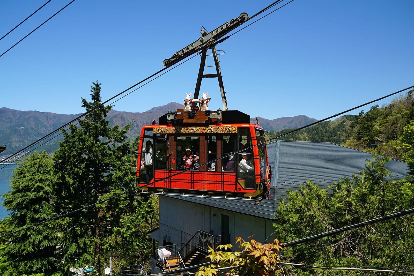 Red Mt. Fuji Panoramic Ropeway gondola with rabbit figures on its roof above the lakeside station, with Lake Kawaguchiko and mountains behind