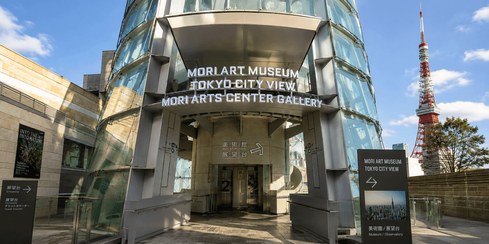 Mori Art Museum entrance signage atop Mori Tower, with Tokyo City View and Mori Arts Center Gallery signage alongside it and Tokyo Tower visible through the glass