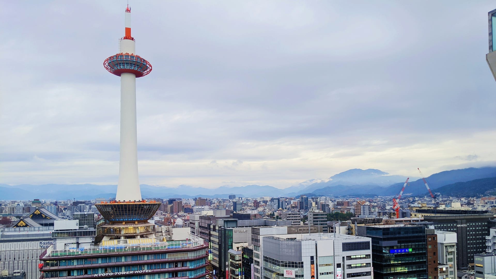Kyoto Tower above the Kyoto Tower Hotel building, with the city and mountains behind on an overcast day