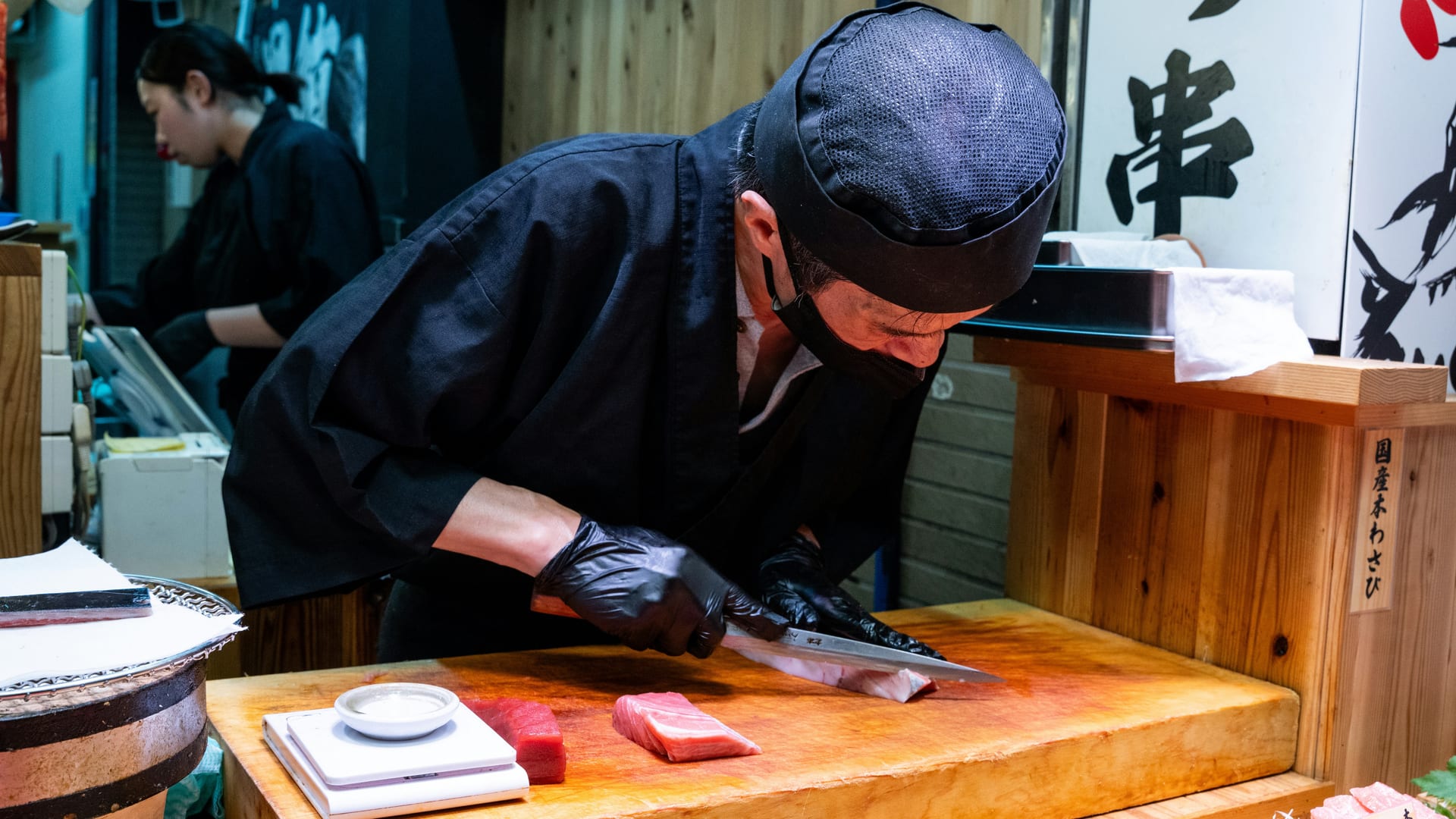 A cook in a black jacket and cap slicing tuna on a wooden board at a fish counter
