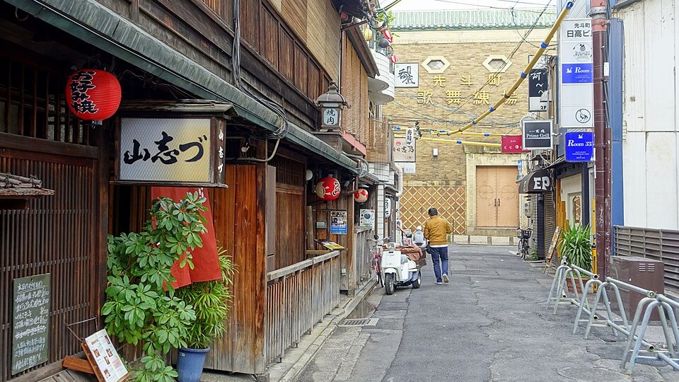 A side lane at the north end of Pontocho by day, with a red lantern and restaurant signs, and the brick Pontocho Kaburenjo theater at the end