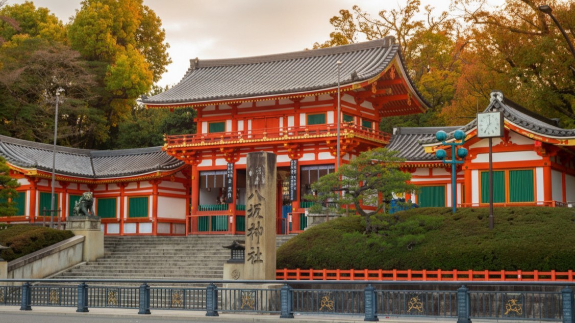 Yasaka Shrine's vermilion West Gate at the top of its stone steps, with the stone name pillar in front and autumn trees behind